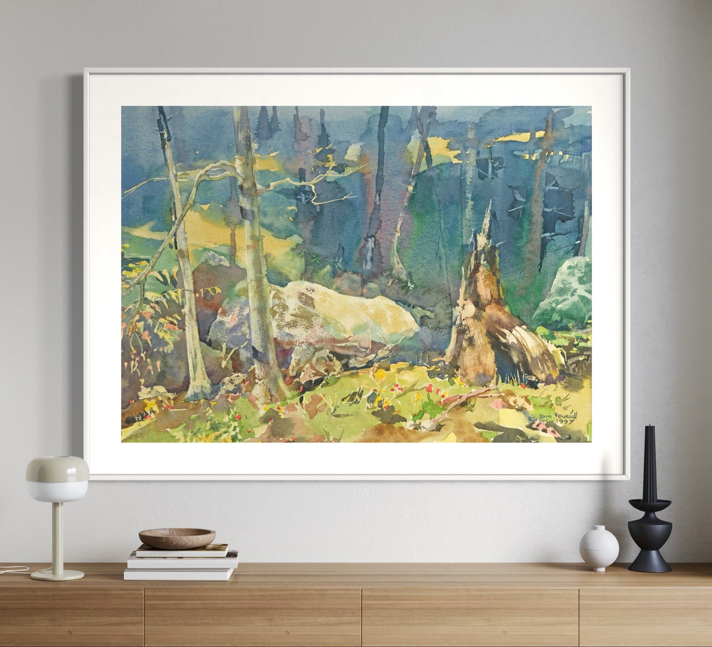 A framed watercolor painting of a wilderness scene with green and yellow colors, displayed on a wooden console table against a white wall.