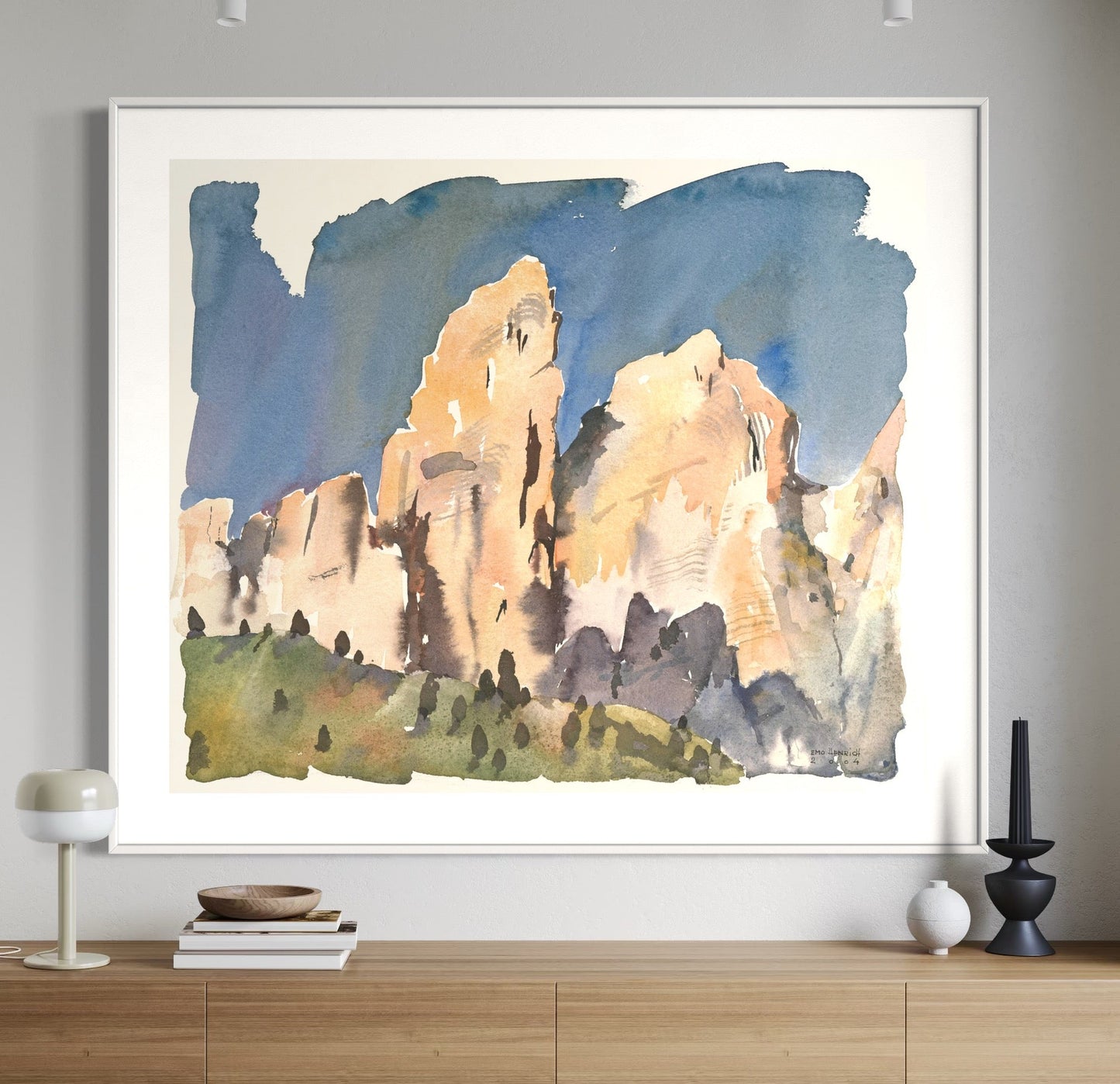 A watercolor painting of a mountainous landscape, titled 'Murfreid-Turm II, Sella, Dolomiten', framed and displayed on a wall above a wooden side table with a lamp and a book.