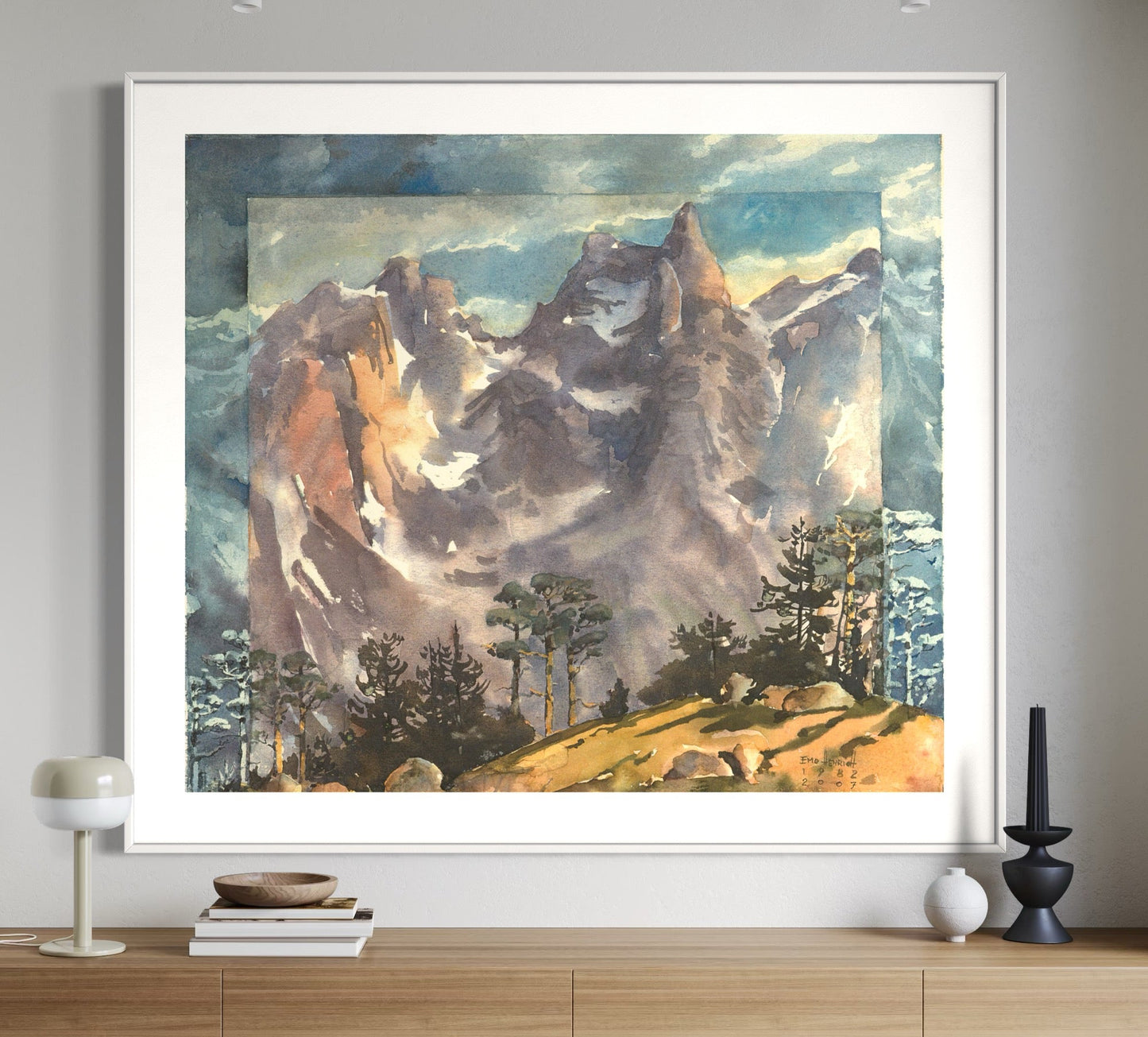 A framed watercolor painting depicting a mountainous landscape with trees in the foreground, displayed on a wooden console table against a white wall.