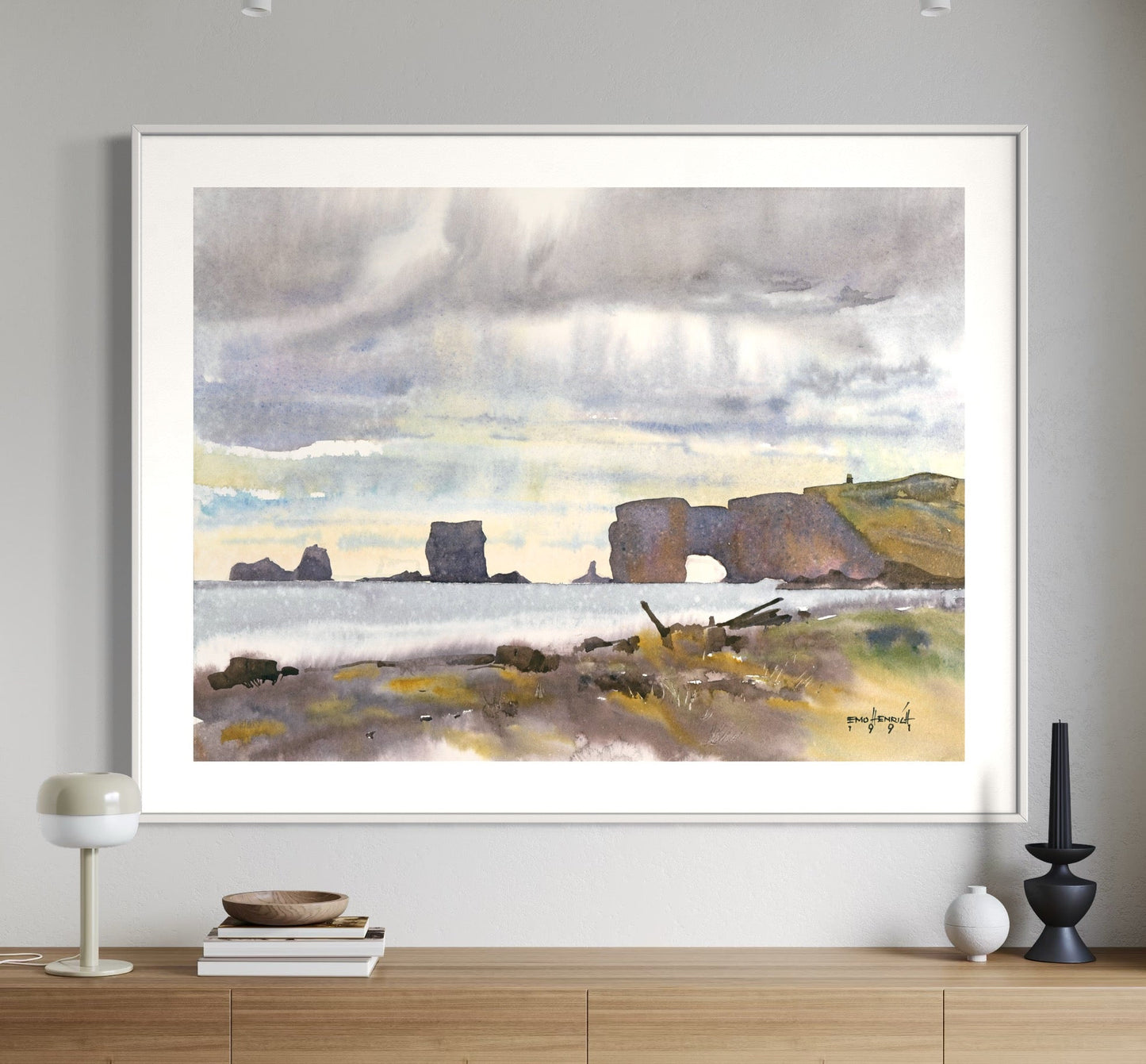 A framed watercolor painting of a cliffside scene, with a large door in the foreground, hanging on a white wall above a side table and chairs.