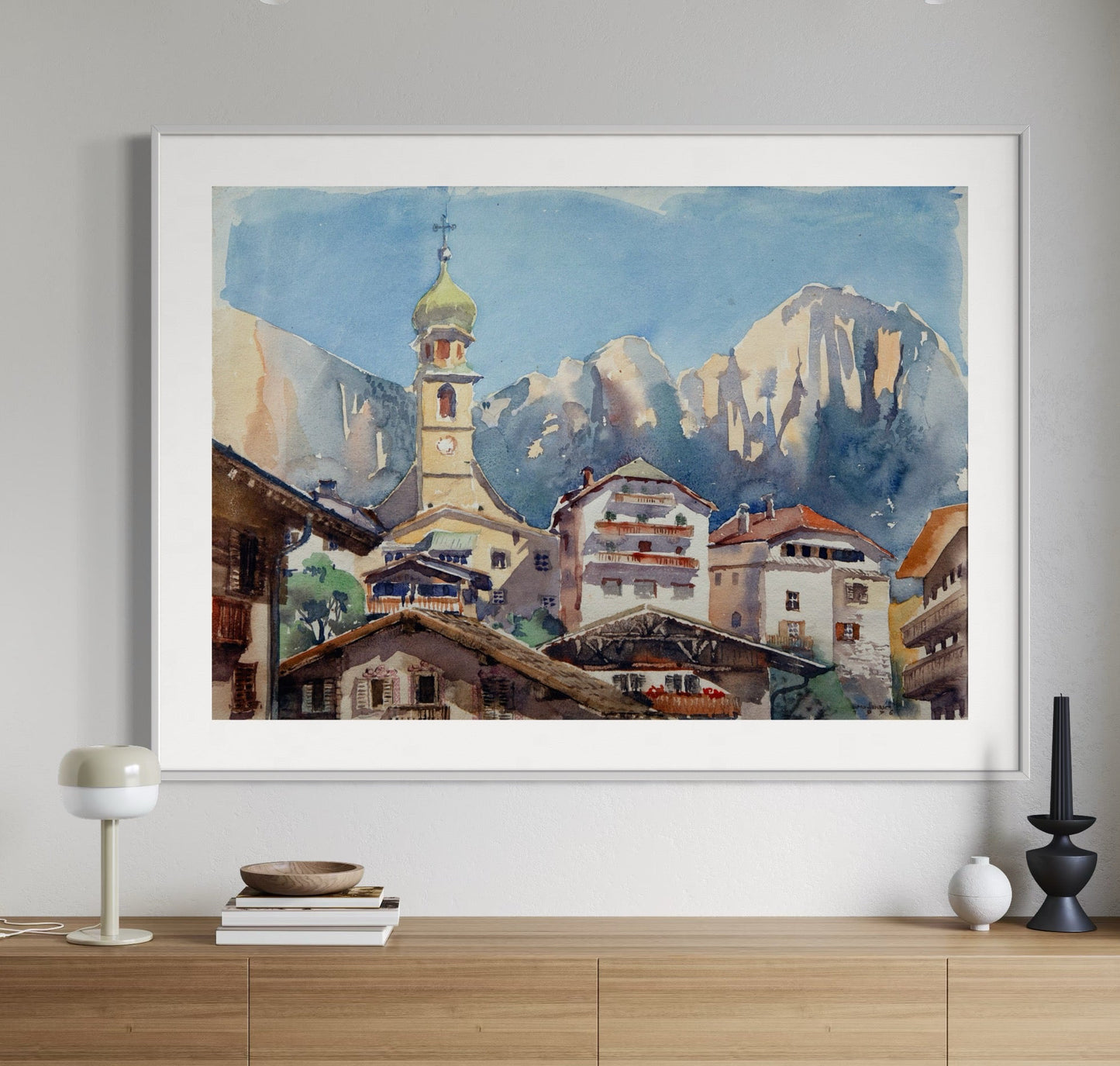 A framed painting of a small town in South Tyrol, Italy, featuring a church and mountains in the background, displayed on a wooden console table.