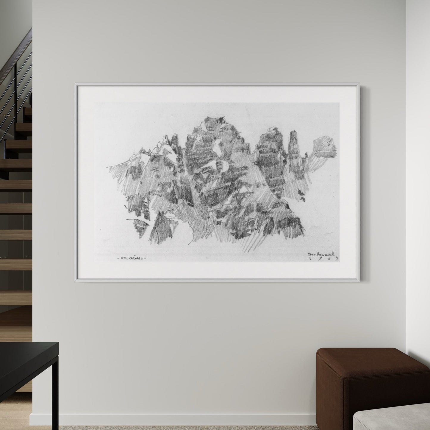 A framed and matted print of a graphite drawing depicting a mountain range, hung on a wall in a room with a modern decor.