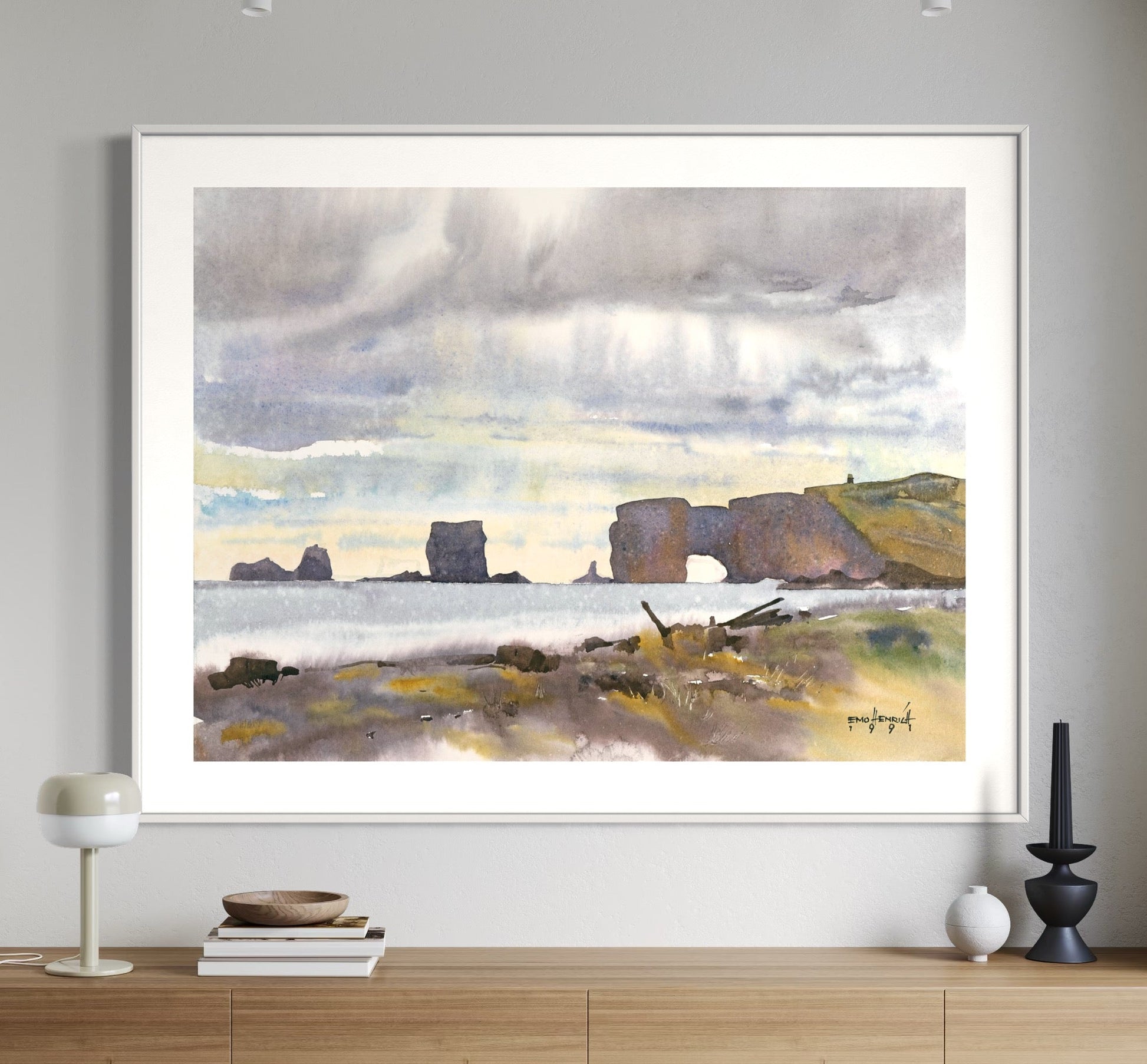 A framed watercolor painting of a cliffside scene, with a large door in the foreground, hanging on a white wall above a side table and chairs.